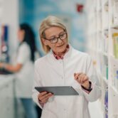 woman-with-tablet-in-pharmacy-aisle-2026-03-25-02-54-34-utc Kobieta z tabletem w alejce apteki sprawdzająca dane o lekach