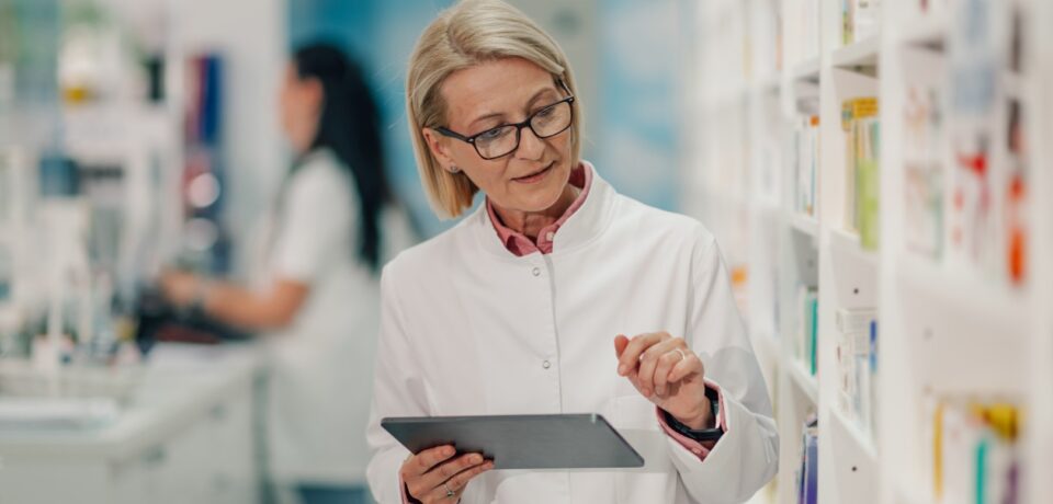 woman-with-tablet-in-pharmacy-aisle-2026-03-25-02-54-34-utc Kobieta z tabletem w alejce apteki sprawdzająca dane o lekach